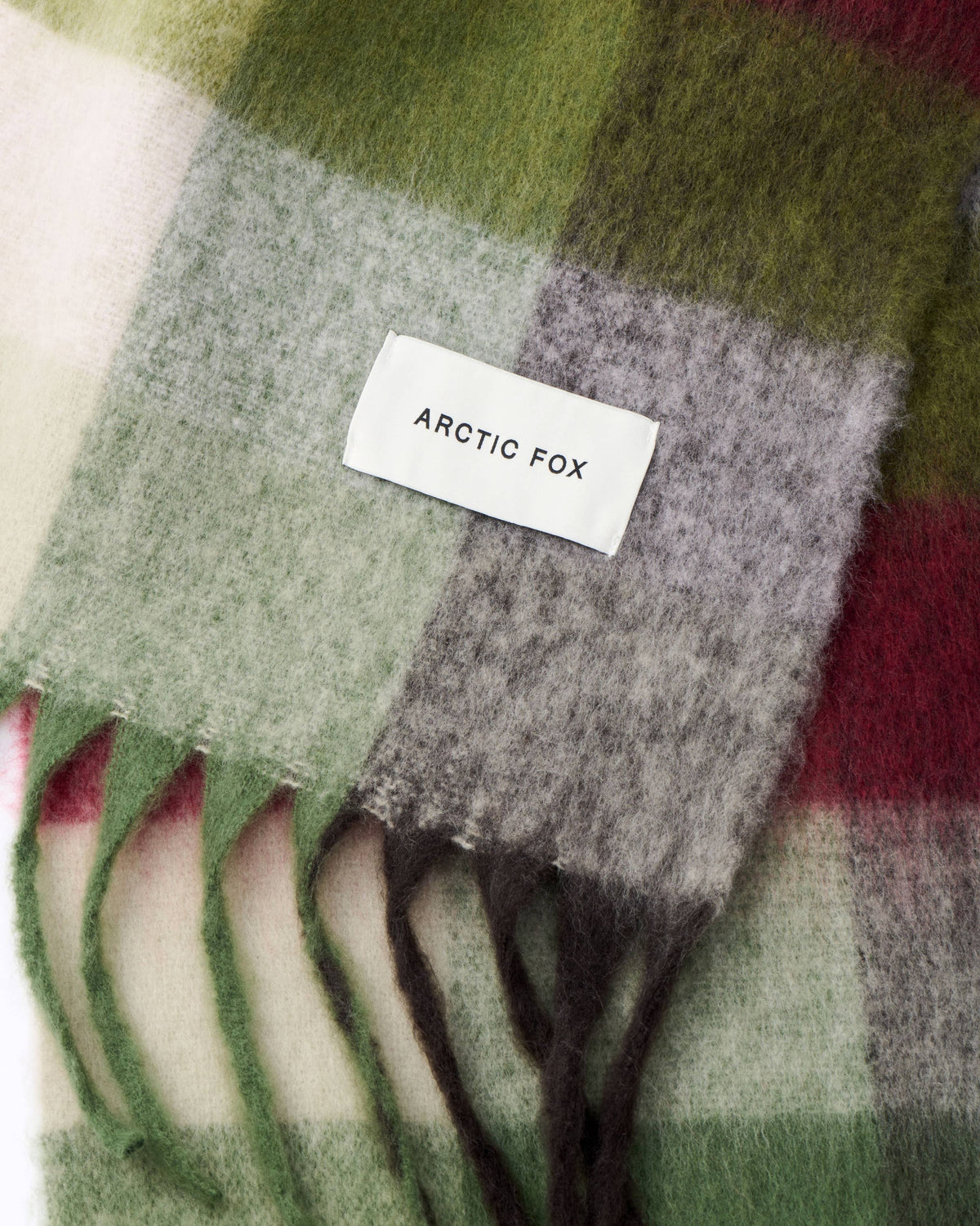 A close-up of a soft plaid scarf with fringe, featuring blocks of sage green, light gray, cream, and deep red. A white woven label reading 'ARCTIC FOX' is stitched onto the scarf.