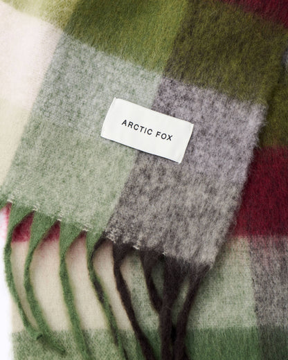 A close-up of a soft plaid scarf with fringe, featuring blocks of sage green, light gray, cream, and deep red. A white woven label reading 'ARCTIC FOX' is stitched onto the scarf.