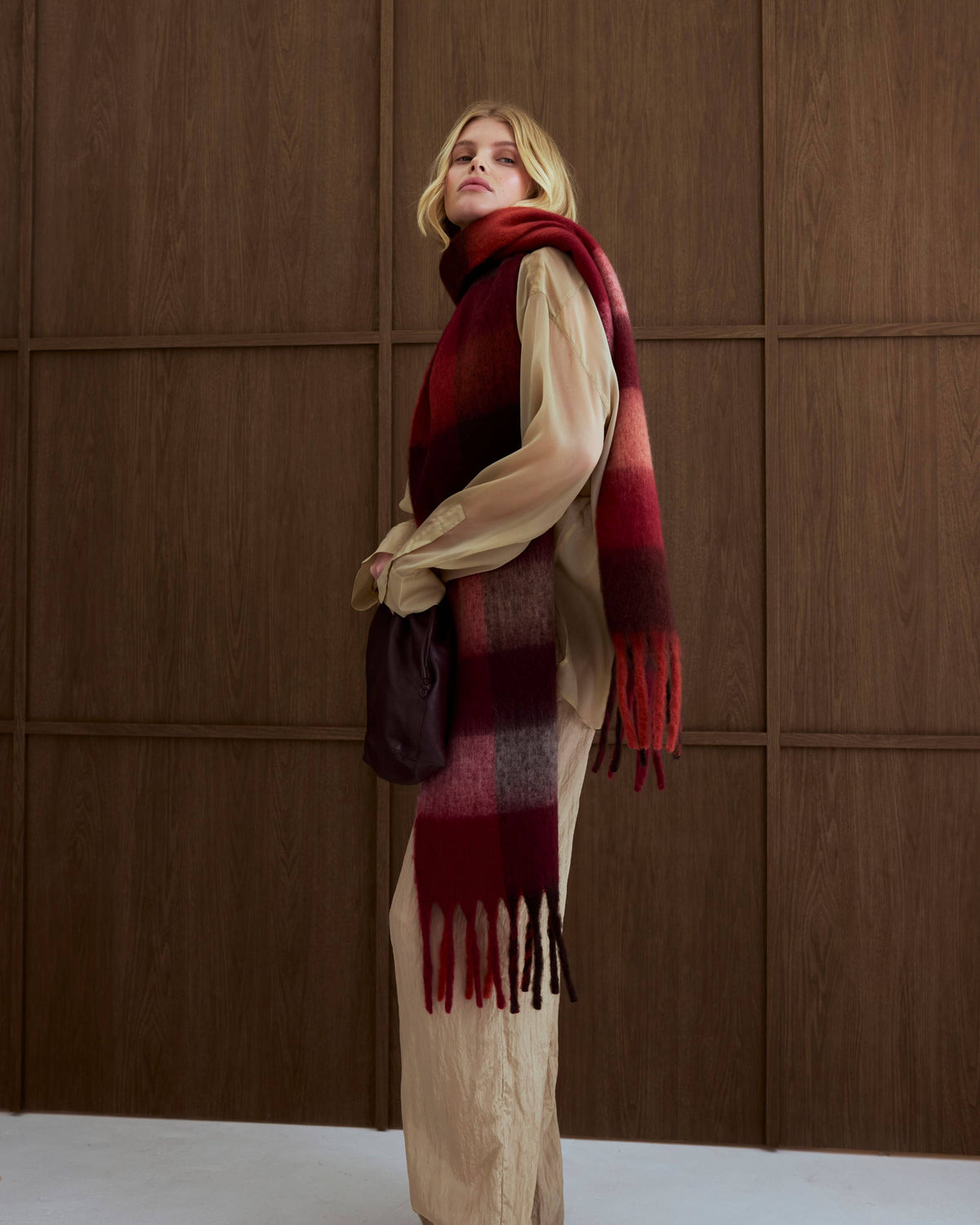 A woman stands in profile wearing a long cream-colored coat and a thick red and dark brown plaid scarf with fringe, holding a dark brown handbag against a brown wood-paneled wall.