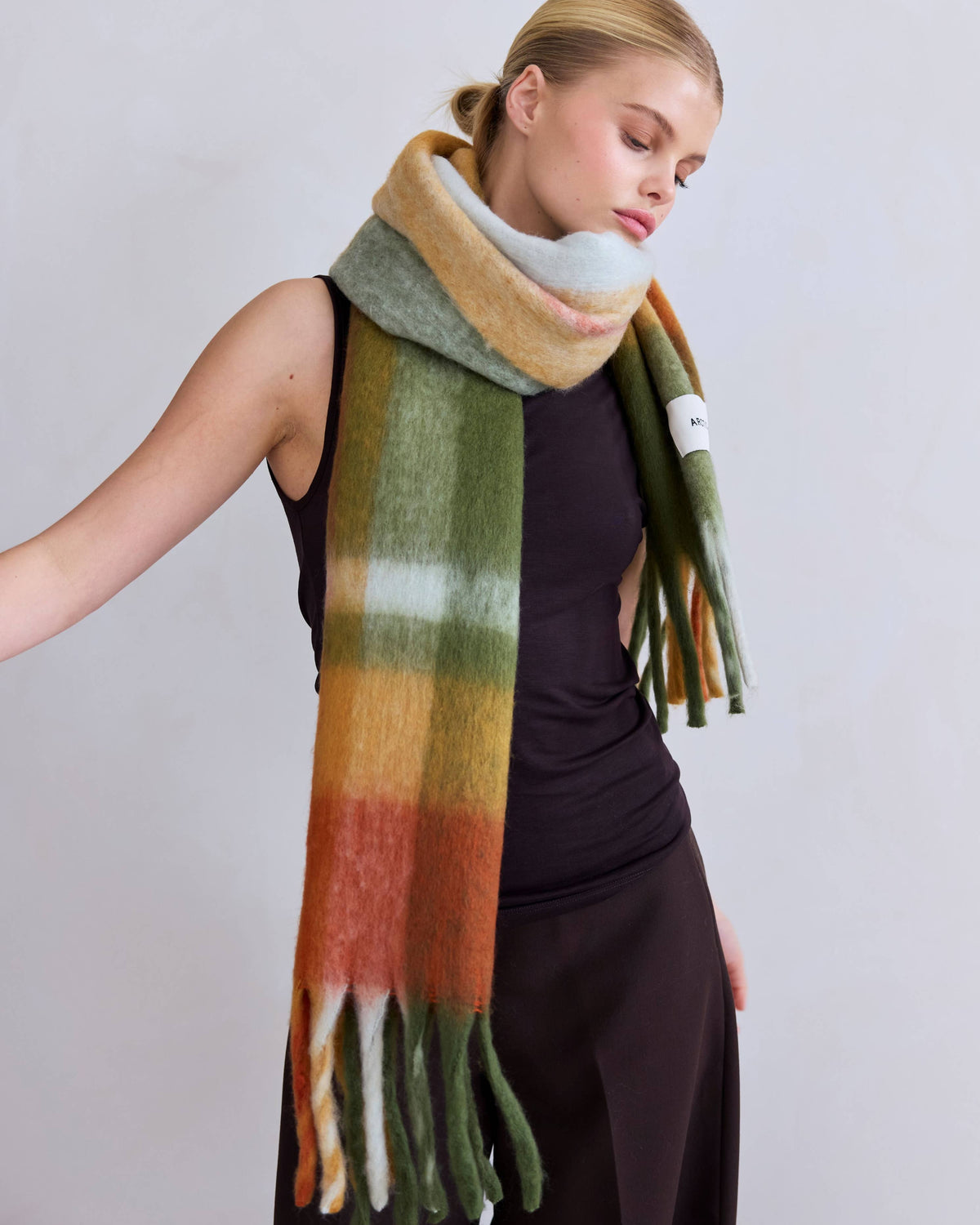 A woman wearing a thick, soft knit scarf with horizontal stripes in sage green, mustard yellow, orange, and cream, styled over a dark brown dress against a light gray studio background.