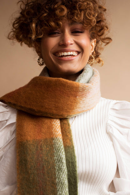 A smiling woman with curly brown hair wearing a thick, soft scarf with a warm gradient of orange, beige, and sage green, paired with a white pleated top against a solid beige background.