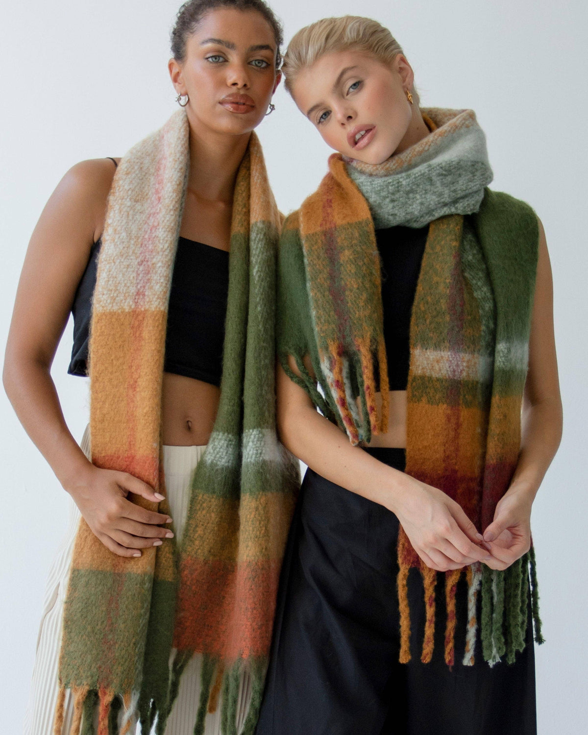 Two models wearing chunky knit fringed scarves. The scarf on the left is a warm beige, orange, and green block pattern, while the scarf on the right is a muted sage green.