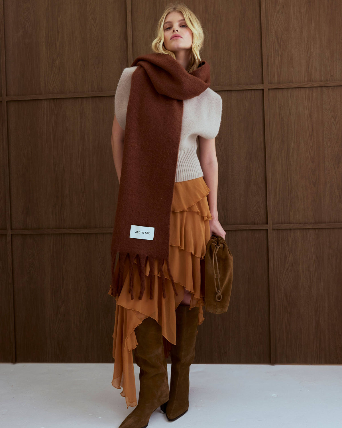 A model stands against a dark wood-paneled wall wearing a cream ribbed knit top, a layered rust-orange ruffled skirt, brown suede boots, and a long brown scarf with a small white label.