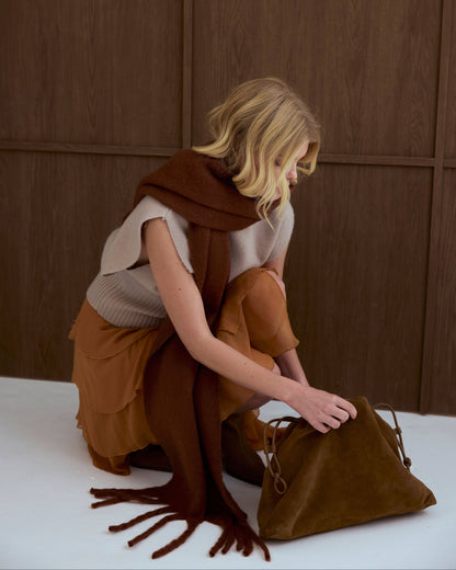 A woman in a beige top and brown skirt sits on a white floor against a dark wood-paneled wall, holding a brown suede-like drawstring bag next to her.