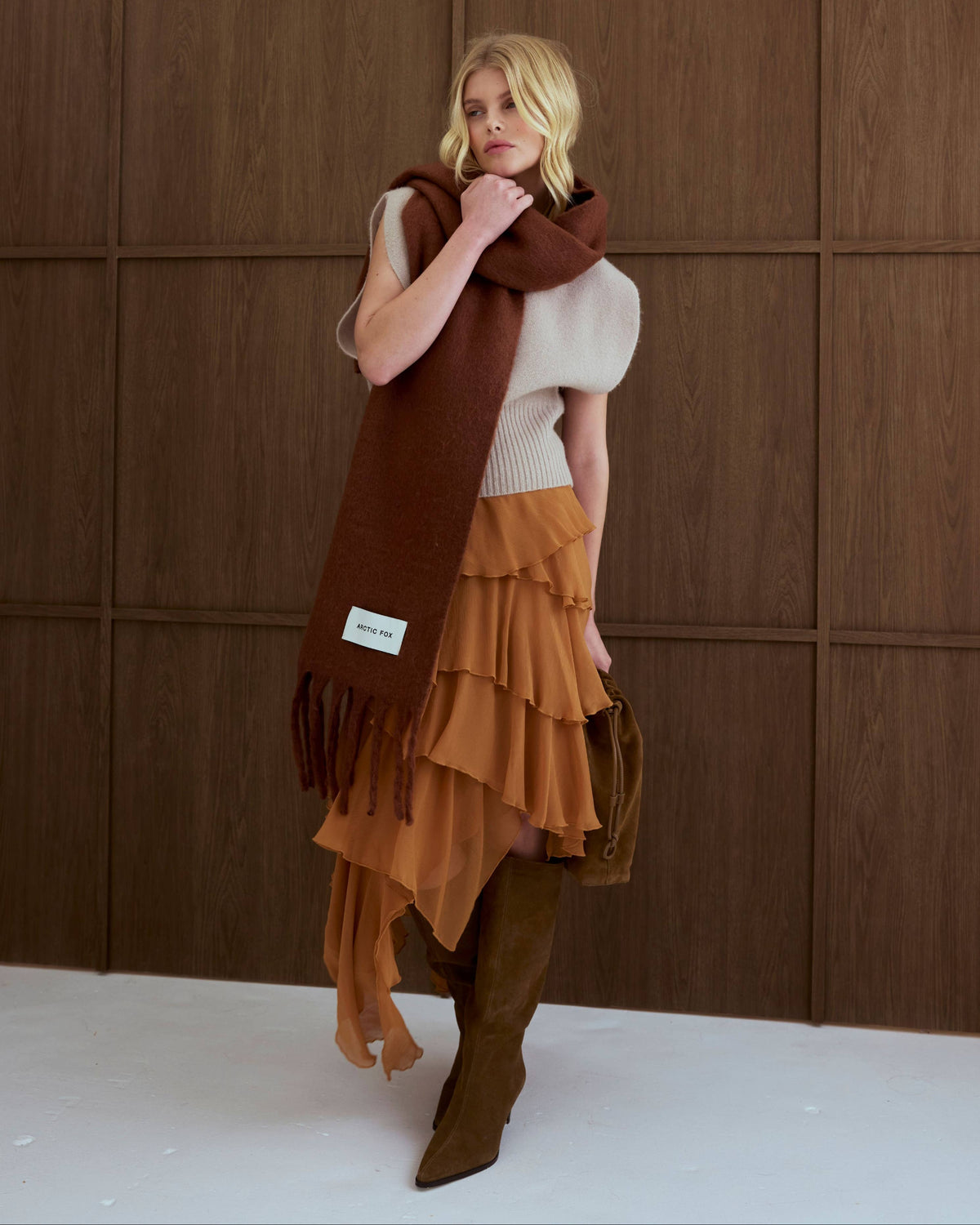 A woman models a stylish autumn outfit featuring a rust-brown ruffled skirt, a cream puff-sleeve top, a dark brown knit scarf, and brown suede boots, standing against a dark wood-paneled wall.