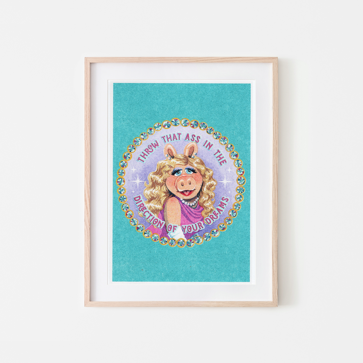 A framed art print featuring a colorful circular illustration of Miss Piggy with the text 'THROW THAT ASS IN THE DIRECTION OF YOUR DREAMS' on a turquoise background, displayed in a light wood frame against a white wall.