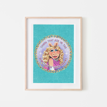 A framed art print featuring a colorful circular illustration of Miss Piggy with the text 'THROW THAT ASS IN THE DIRECTION OF YOUR DREAMS' on a turquoise background, displayed in a light wood frame against a white wall.