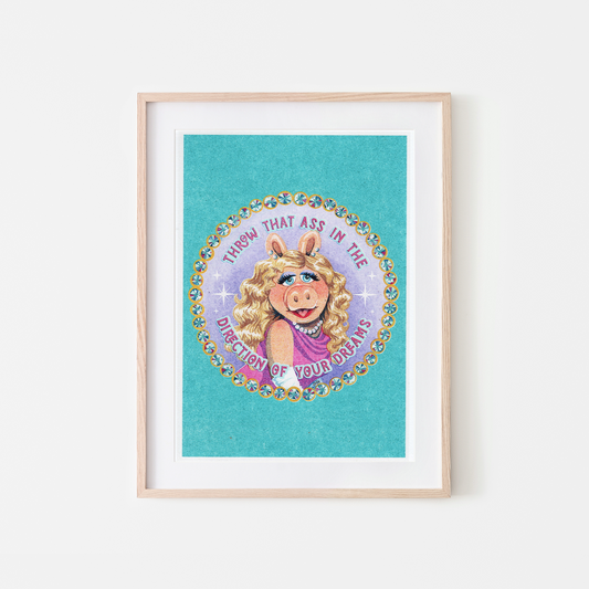 A framed art print featuring a colorful circular illustration of Miss Piggy with the text 'THROW THAT ASS IN THE DIRECTION OF YOUR DREAMS' on a turquoise background, displayed in a light wood frame against a white wall.