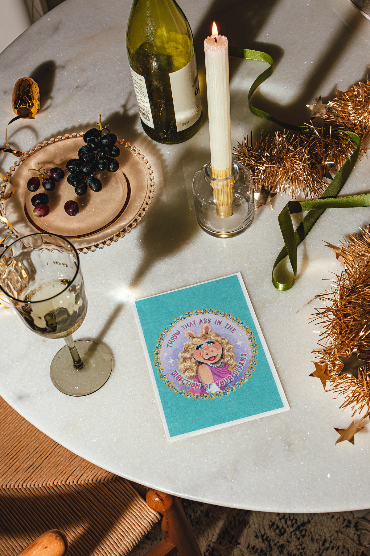 A styled tabletop scene with a turquoise greeting card featuring MISS PIGGY and the text 'THROW THAT ASS IN THE DIRECTION OF YOUR DREAMS', alongside a wine bottle, lit candle, glass, grapes on a plate, ribbons, and gold tinsel on a light marble surface.