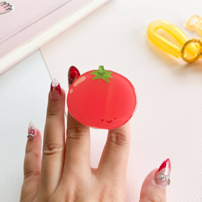 Hand holding a red tomato-shaped compact mirror with a green stem on a light pink background.