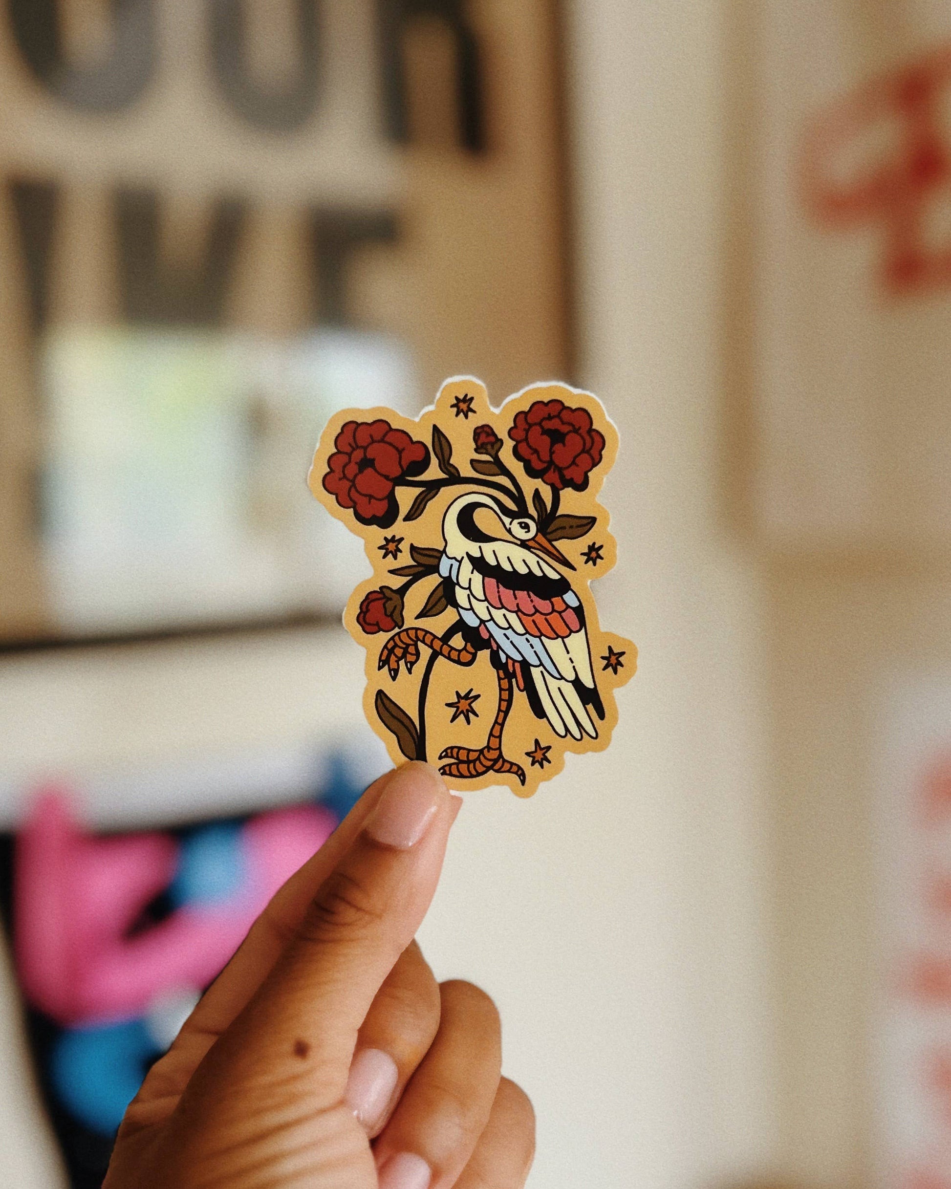 A hand holds a small die-cut sticker featuring a traditional tattoo-style bird with red flowers and a blue-and-white striped body, against a softly blurred indoor background with posters.