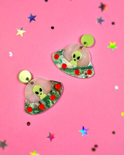 Pair of earrings with alien design on a pink background with star confetti
