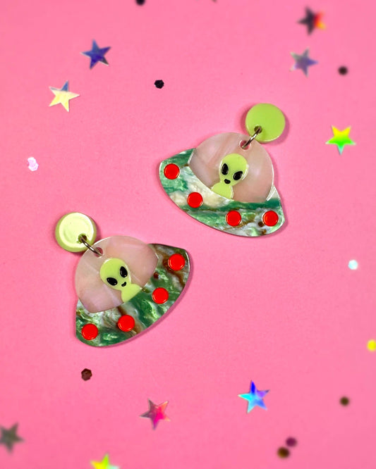 Pair of earrings with alien design on a pink background with star confetti