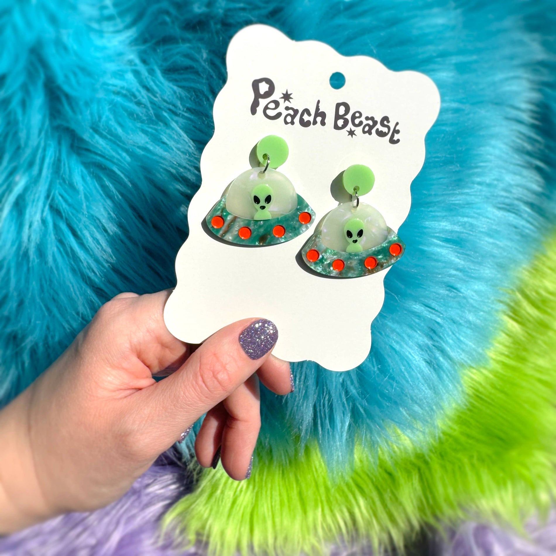 Pair of UFO Alien earrings with a 'Peach Beast' label held against a colorful, fluffy background.