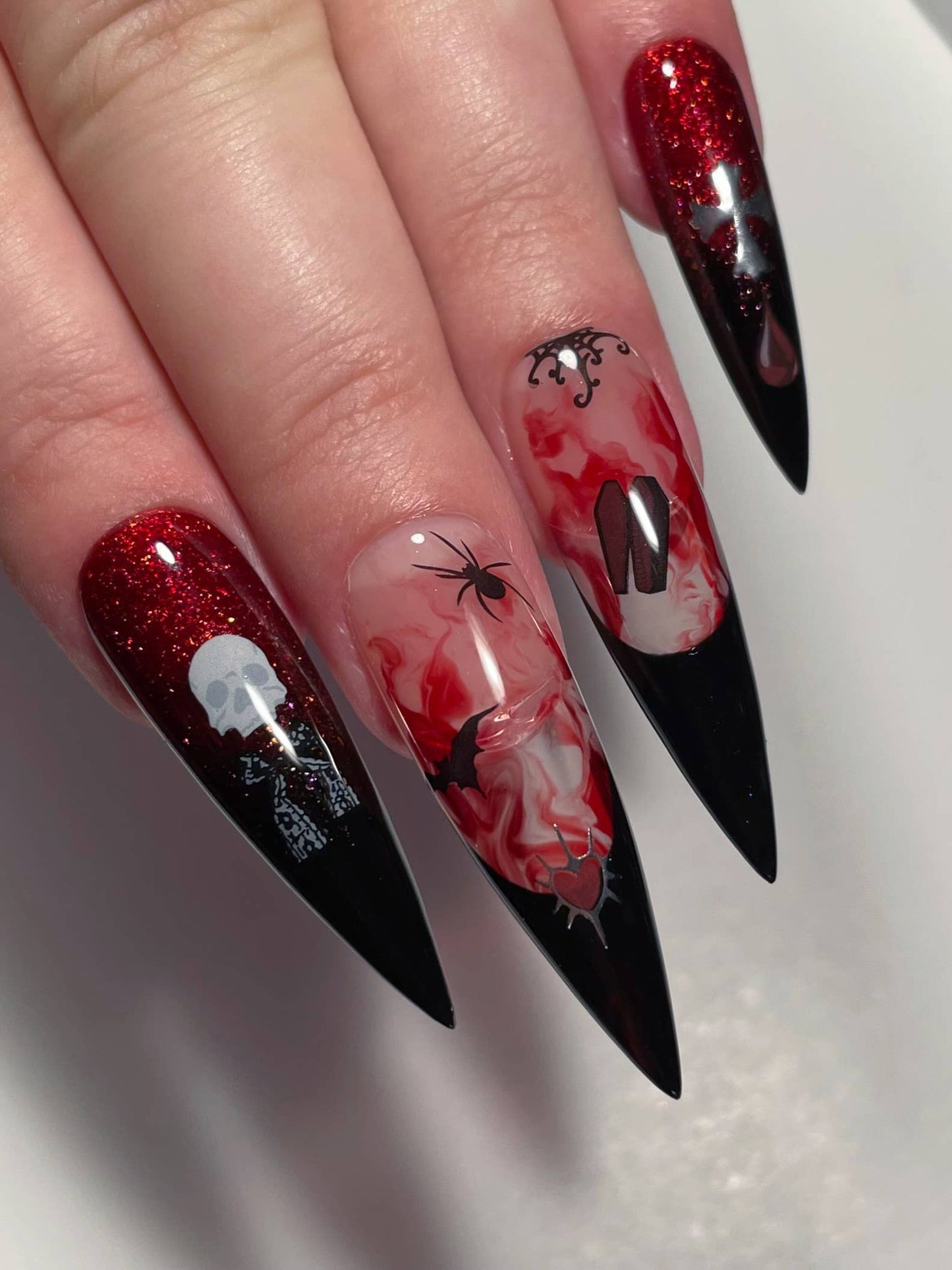 A close-up of a hand with long, pointed stiletto nails featuring a gothic Halloween design. The nails have a black-to-red glitter gradient, with white skull and skeleton details on one nail, a spider and bat on another, and a red-and-white marbled pattern with a black letter 'N' on the third.