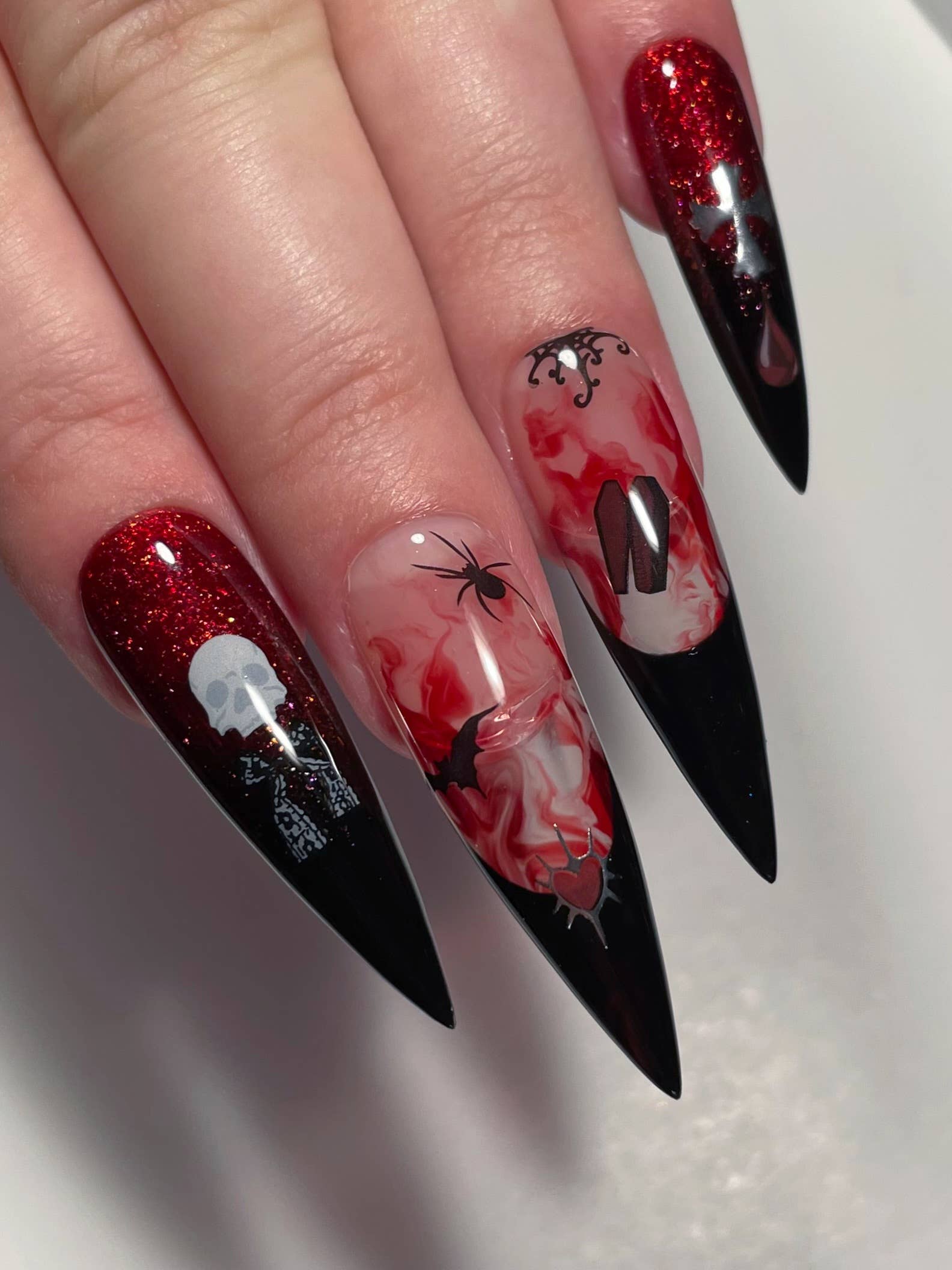 A close-up of a hand with long, pointed stiletto nails featuring a gothic Halloween design. The nails have a black-to-red glitter gradient, with white skull and skeleton details on one nail, a spider and bat on another, and a red-and-white marbled pattern with a black letter 'N' on the third.