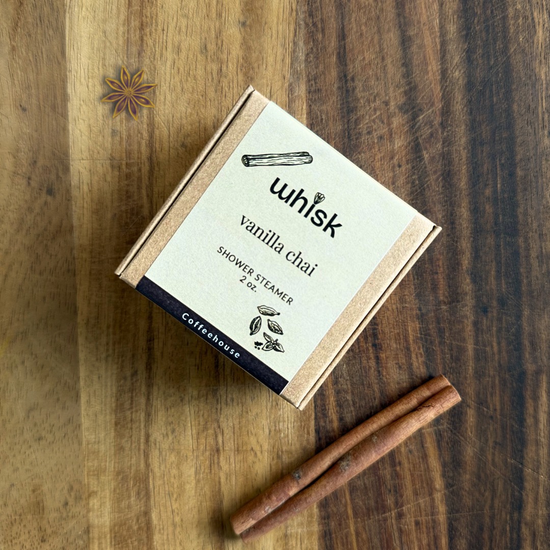 A kraft paper box labeled 'whisk vanilla chai shower steamer 2 oz' by Coffeehouse, placed on a wooden surface with two cinnamon sticks nearby.