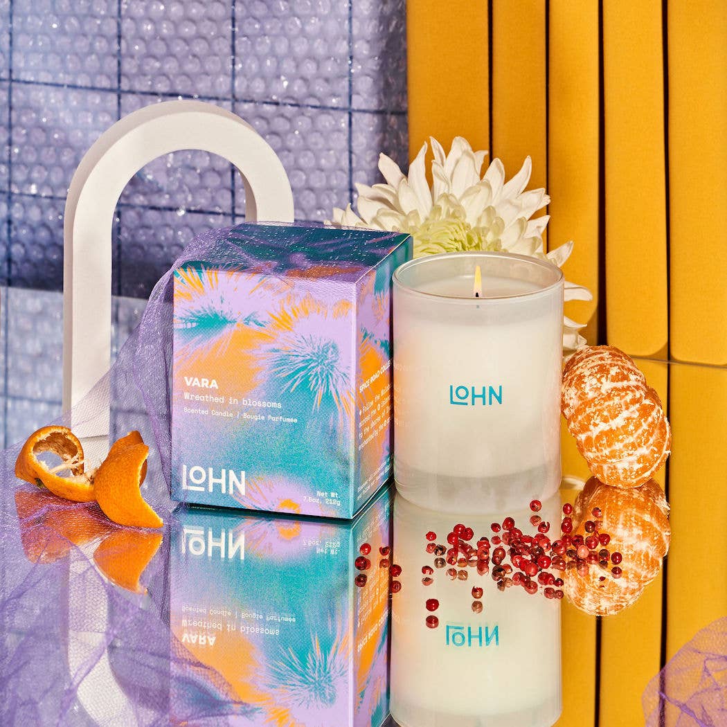 A boxed candle and an unboxed white candle jar with visible LOHN branding, styled on a reflective surface with orange slices, a flower, and a yellow-orange striped backdrop.