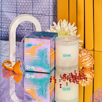A boxed candle and an unboxed white candle jar with visible LOHN branding, styled on a reflective surface with orange slices, a flower, and a yellow-orange striped backdrop.