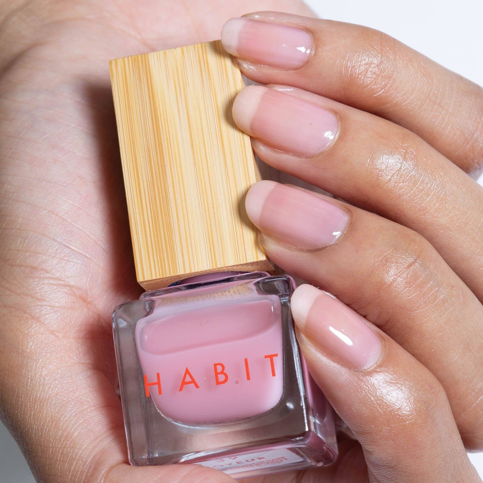 Hand holding a pink nail polish bottle with wooden cap labeled 'Habit'.
