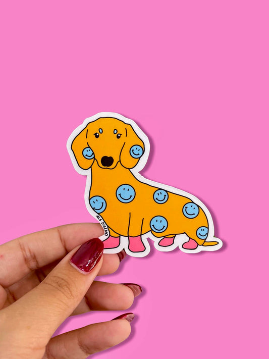 Sticker of a dog with blue smiley face spots held by a hand on a pink background