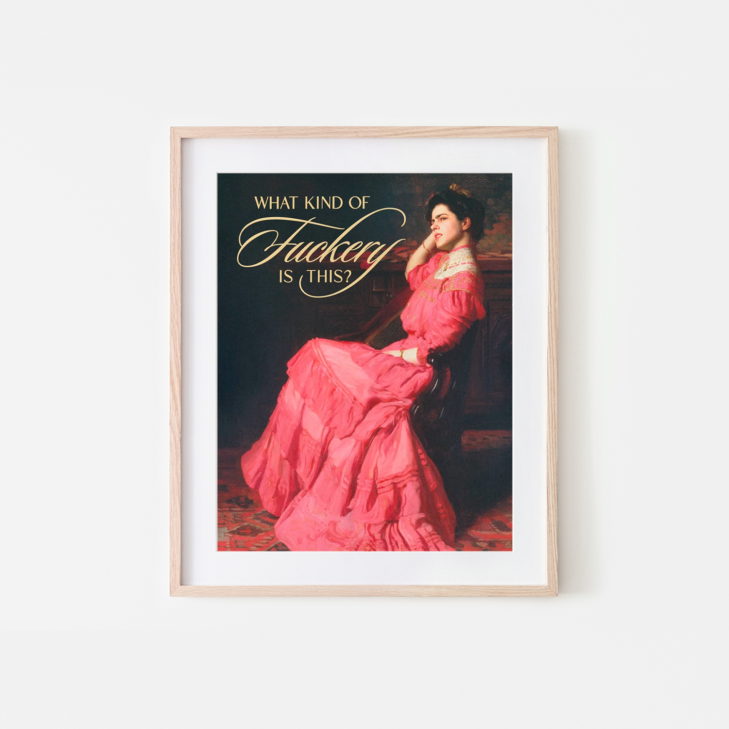 A framed art print featuring a parody of a classic oil painting of a woman in a pink gown, with the text 'WHAT KIND OF F*CKTAY IS THIS?' in gold lettering. The print is displayed in a light wood frame with a white mat against a clean white wall.