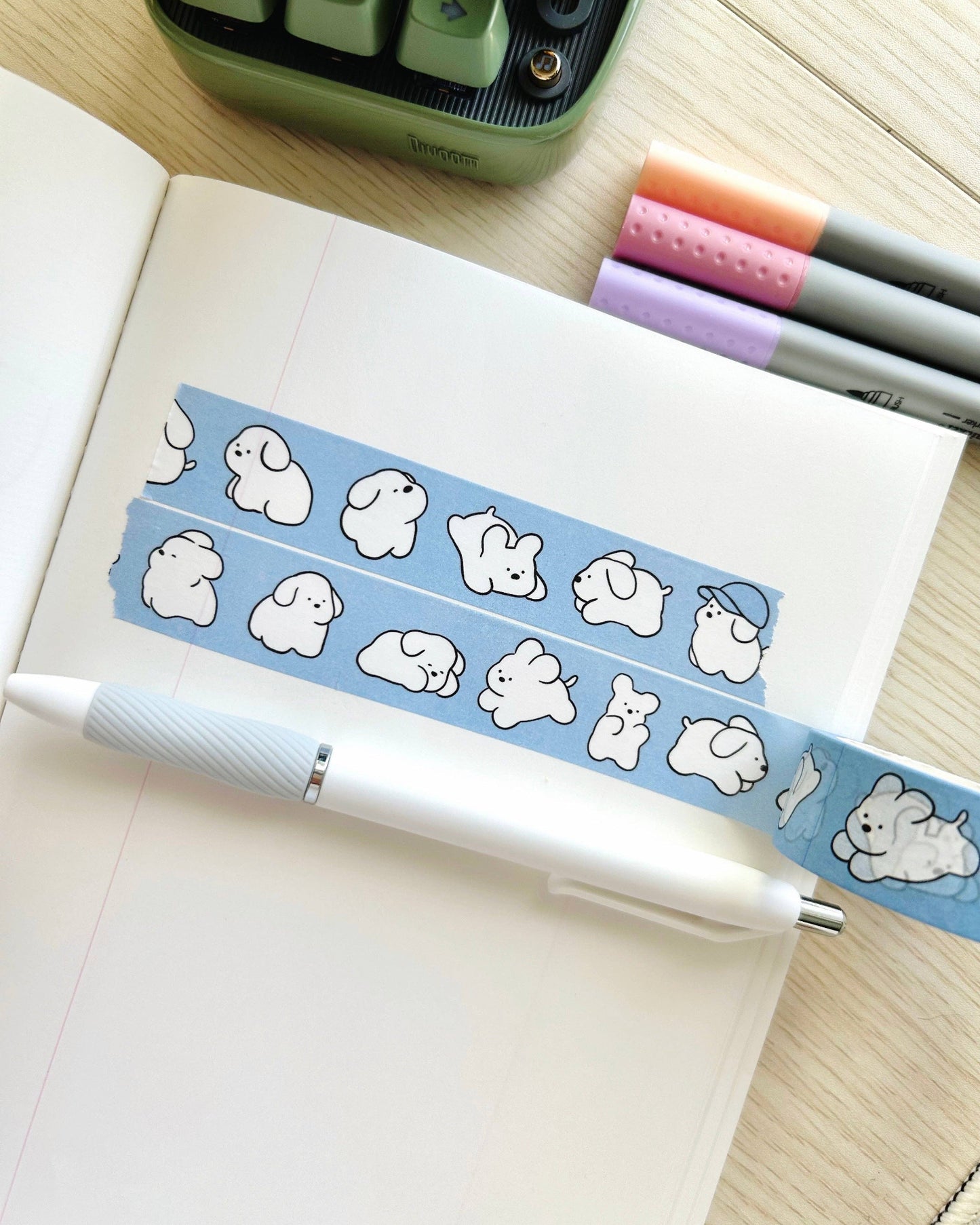 Notebook with a blue washi tape featuring cartoon dog designs, a pen, and stationery items on a wooden surface. White Puppy Washi Tape