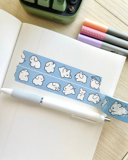 Notebook with a blue washi tape featuring cartoon dog designs, a pen, and stationery items on a wooden surface. White Puppy Washi Tape