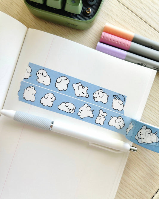 Notebook with a blue washi tape featuring cartoon dog designs, a pen, and stationery items on a wooden surface. White Puppy Washi Tape