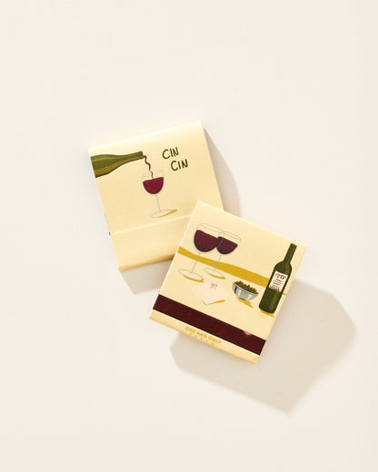 Two square cream-colored matchbook-style coasters with wine-themed illustrations, including a bottle, glasses of red wine, and the word 'CIN CIN', photographed on a light beige background.