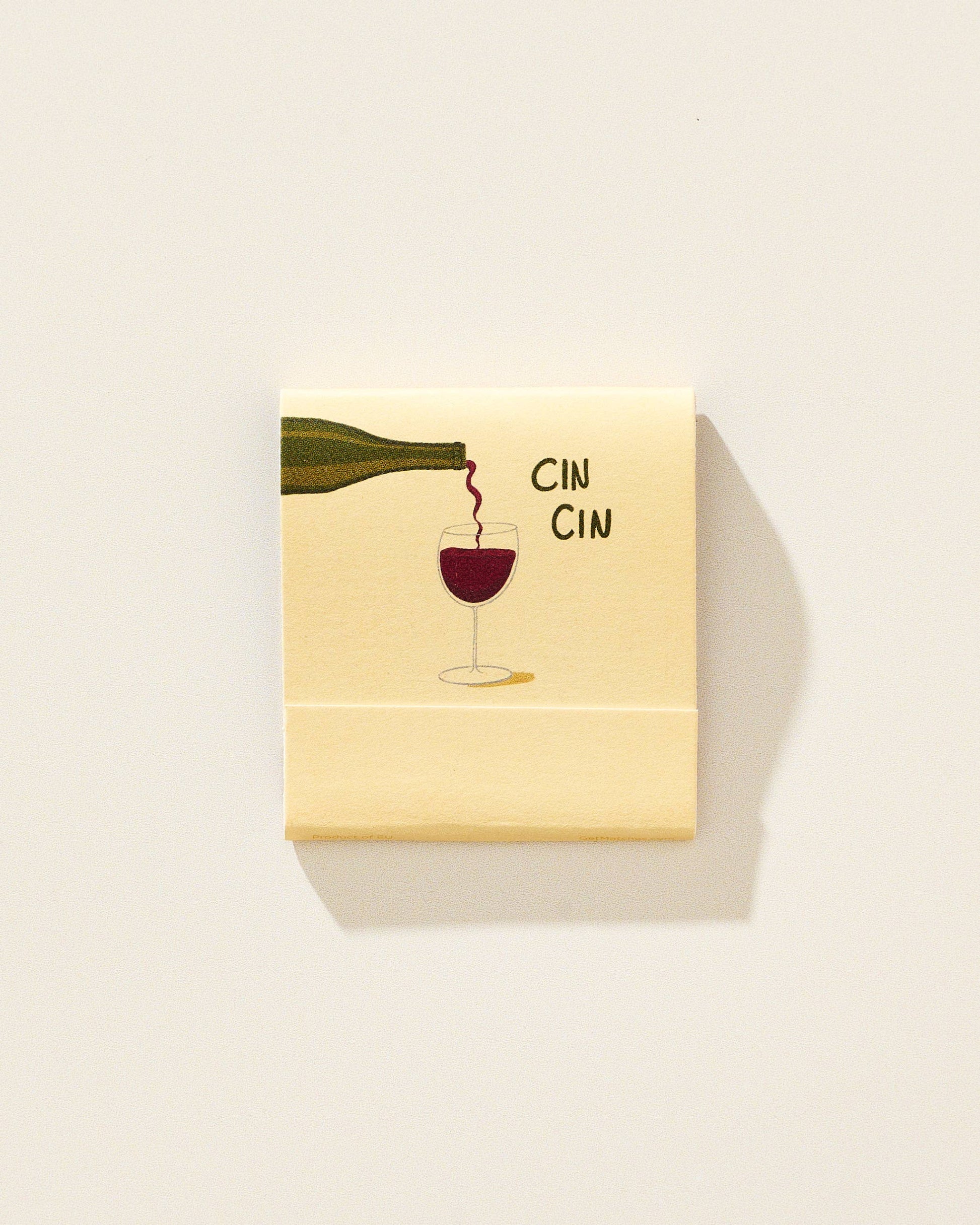 A square cream-colored matchbook with a printed illustration of a wine bottle pouring red wine into a glass, alongside the text 'CIN CIN', shown on a plain light background.