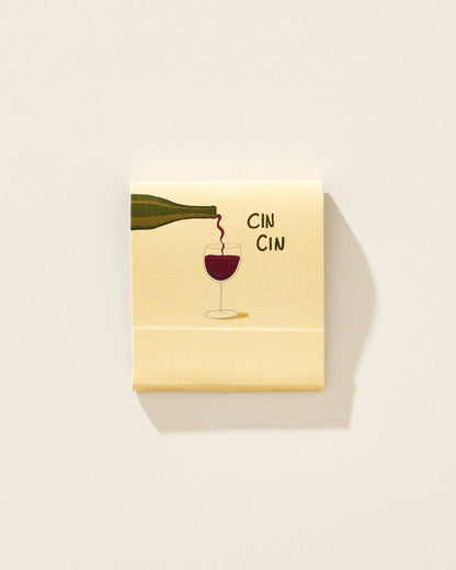 A square cream-colored matchbook with a printed illustration of a wine bottle pouring red wine into a glass, alongside the text 'CIN CIN', shown on a plain light background.