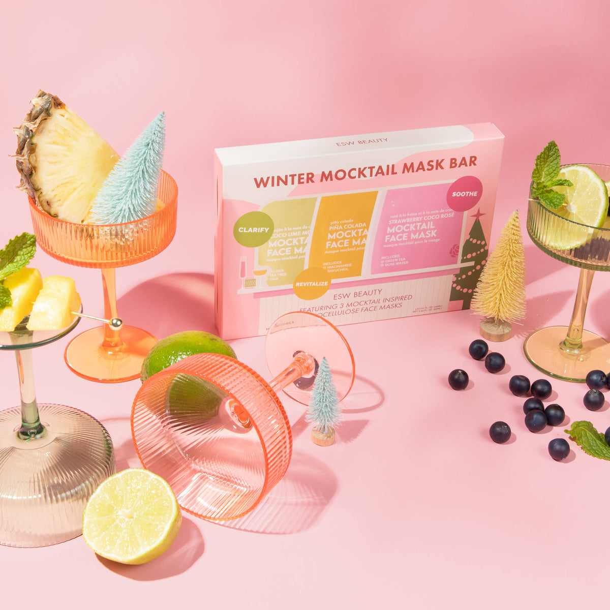 A boxed set called Winter Mocktail Mask Bar sits on a pink studio background, surrounded by cocktail glasses with pineapple, lime, and blueberries, plus small decorative trees and a pink mask bar handle. The box shows CLARIFY and ROOSTE branding and reads WINTER MOCKTAIL MASK BAR.