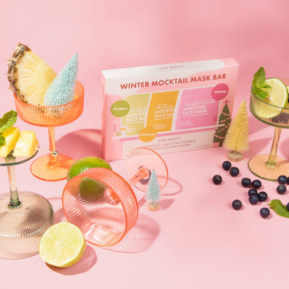A boxed set called Winter Mocktail Mask Bar sits on a pink studio background, surrounded by cocktail glasses with pineapple, lime, and blueberries, plus small decorative trees and a pink mask bar handle. The box shows CLARIFY and ROOSTE branding and reads WINTER MOCKTAIL MASK BAR.