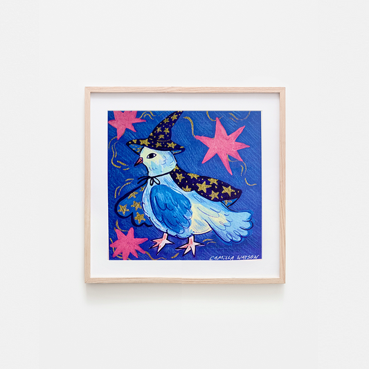 A framed art print featuring a whimsical illustration of a blue bird dressed as a witch with a star-patterned hat and cape, set against a dark blue background with pink starbursts, displayed in a light wood frame on a white wall.